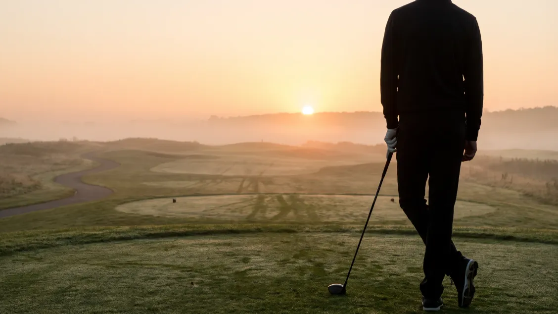 Golfer seen from behind on an empty tee at sunrise, driver held low at one side, mist lying over the fairway ahead