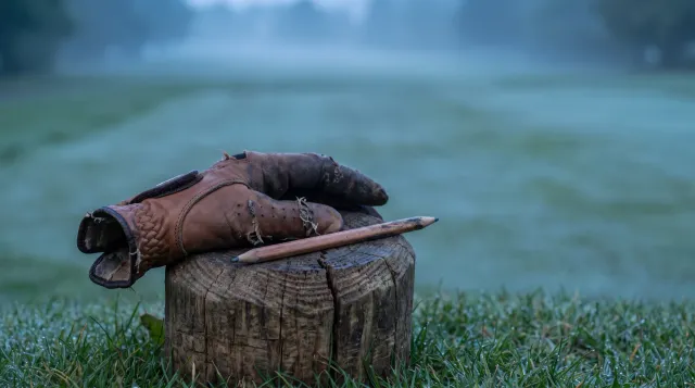 Weathered leather golf glove and a pencil resting on a cut wooden post in dewy grass, empty fairway soft and out of focus behind in blue pre-dawn light
