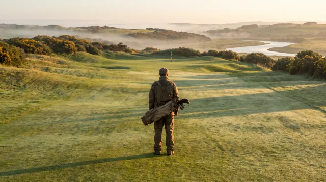 Lone golfer standing on a misty links fairway at sunrise, carrying his bag, looking down the hole ahead