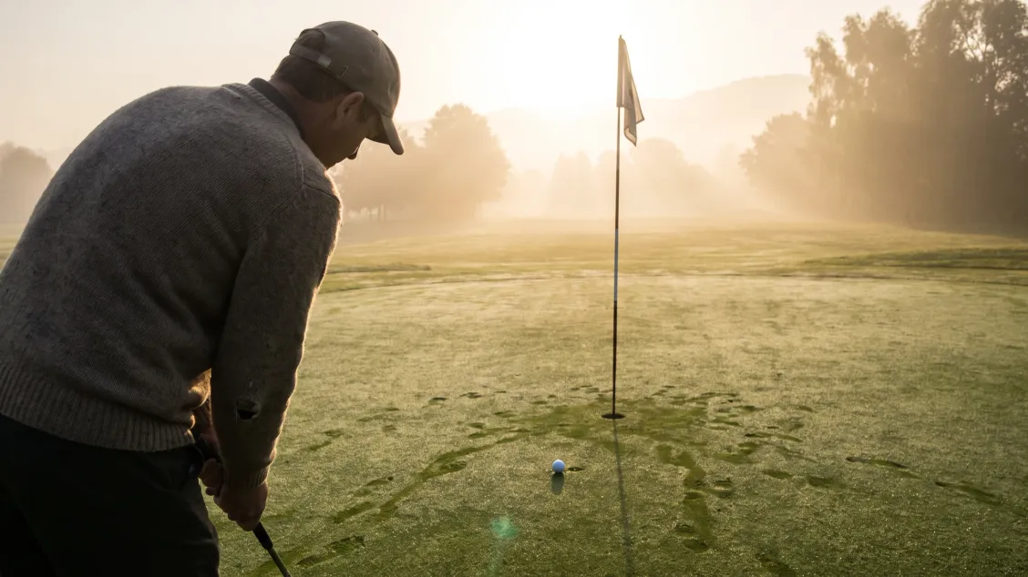 Golfer in a grey sweater and cap standing over a short putt on a misty green at sunrise, flagstick in the hole a few feet ahead