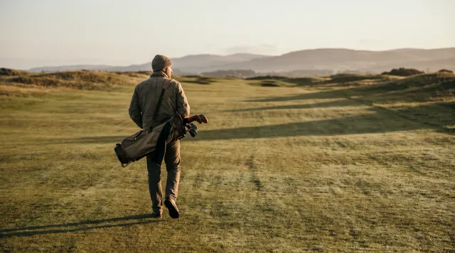 Golfer walking up a quiet fairway with his head up and his bag on his shoulder, soft morning light and long shadows