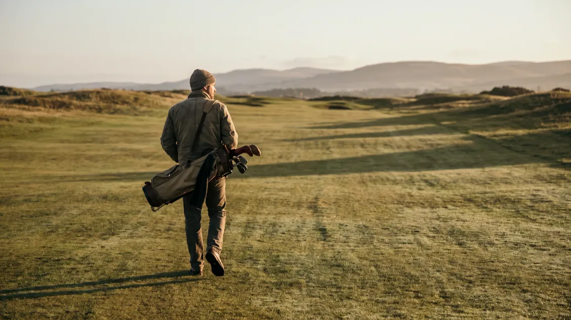 Golfer walking up a quiet fairway with his head up and his bag on his shoulder, soft morning light and long shadows