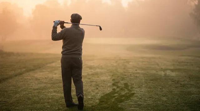 Golfer seen from behind walking up a misty fairway at dawn, iron resting across his shoulders, empty course ahead of him
