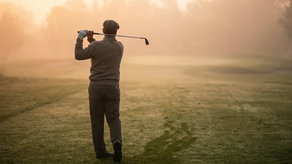 Golfer seen from behind walking up a misty fairway at dawn, iron resting across his shoulders, empty course ahead of him