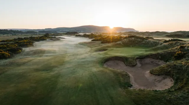 Sunrise over a links hole seen from above, a wide open fairway sweeping left through low mist and a small green tucked right behind three deep bunkers